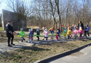 Dzieci wraz z paniami maszerują z wężem ulicami osiedla, w ręku trzymają papierowe kwiatki, śpiewają wiosenne piosenki.